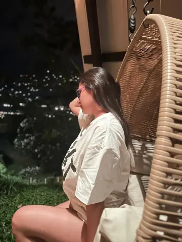 Young woman in a white t-shirt relaxing in a wicker hanging chair at night in a lush garden setting.