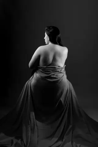 Elegant woman in a flowing draped gown posing from behind in a moody black and white studio portrait.