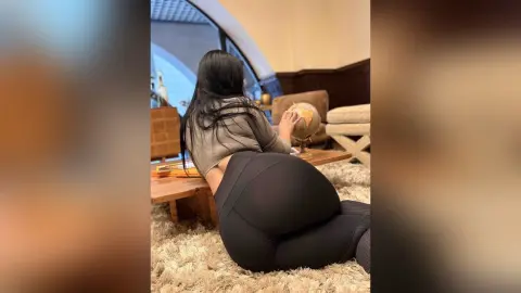 JasminNise shows off her large, rounded butt in tight black leggings while leaning over a wooden coffee table in a living room setting.
