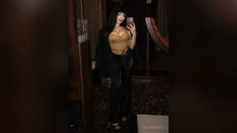 Glamorous woman in a gold corset and black velvet trousers taking a mirror selfie in a moody lounge setting.