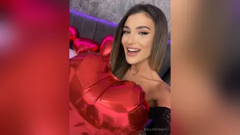 Smiling brunette woman in a black lace outfit holding red heart balloons in a cozy room.