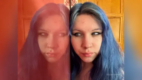 DeidreSarin poses with her long blue hair against a wooden door, looking off to the side with heavy eyeliner.