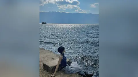 Relaxed woman in a blue tank top sitting by a sparkling lake with mountains under a blue sky.