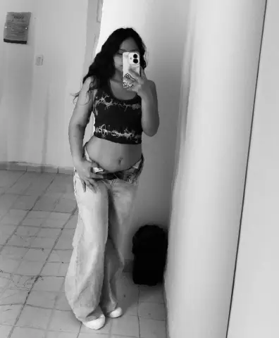 Young woman in a patterned tank top and baggy jeans taking a mirror selfie in a minimalist room.