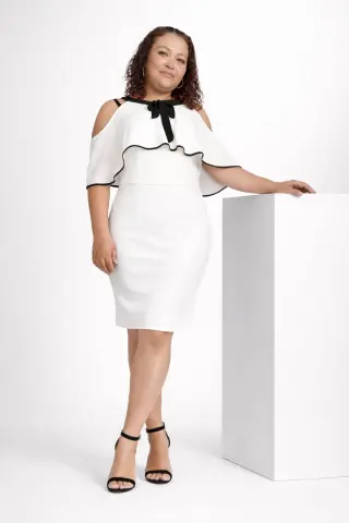 Smiling woman in a white sleeveless cocktail dress with black trim standing against a white studio background.