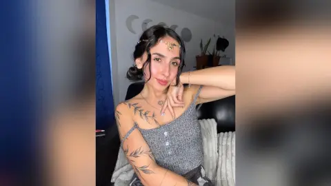 RossiNoleen poses in a silver glittery tank top, showing off her dark hair and leaf tattoos on her shoulder and arm.
