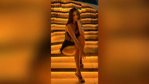 TefyRusso sits on lit stairs wearing a black bodysuit and high heels, posing with her long dark hair down.