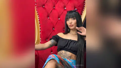 Stylish woman with long black hair and tattoos wearing a black crop top on a red velvet throne.