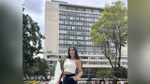 Stylish woman in a white crop top and black trousers standing by a city building and green trees.