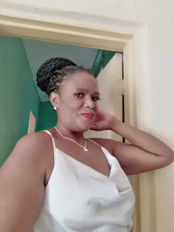 Smiling woman in a white camisole tank top posing indoors against a teal wall.