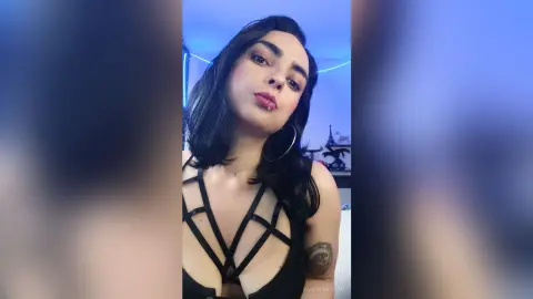 Young woman with long dark hair wearing a black strappy cutout bodysuit in a moody blue-lit room.