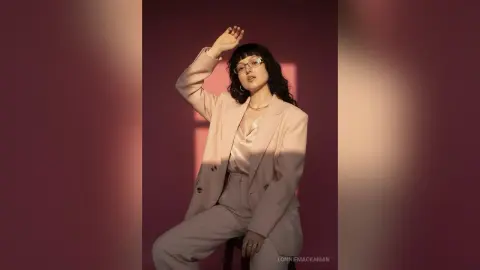 Stylish woman in a beige blazer and trousers posing against a warm pink background with soft lighting.