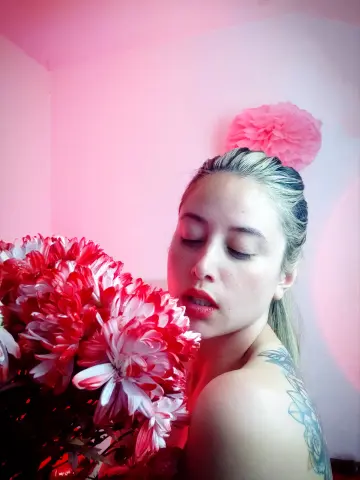 Blonde woman with tattoos holding red and white flowers in a dreamy pink lit room.