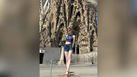 Young woman in a navy star crop top and denim shorts standing before the Sagrada Familia in sunny Barcelona.