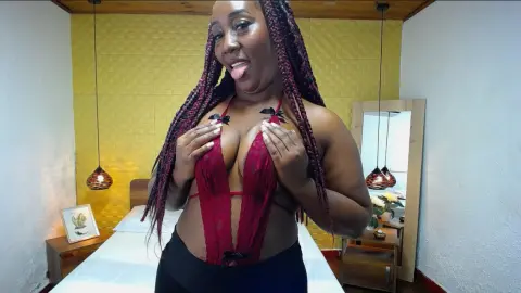 Smiling woman with long braids wearing a red lace bodysuit in a bright bedroom setting.