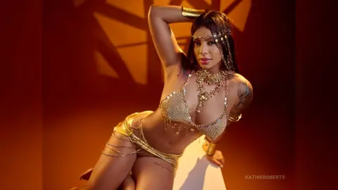 Glamorous woman in gold sequin bikini and jewelry posing in warm, sunlit studio setting.