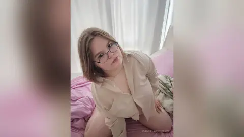 ManeelaKennedy wears glasses and an open beige shirt while sitting on a pink bed.