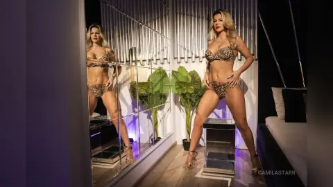 CamilaStarr poses in a leopard print bikini, standing in a room with a large mirror, green plants, and a bed behind her.