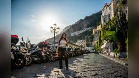 MissyJolie stands on a stone street wearing a white tank top and black patterned leggings, with a bright sunlit hillside behind her.