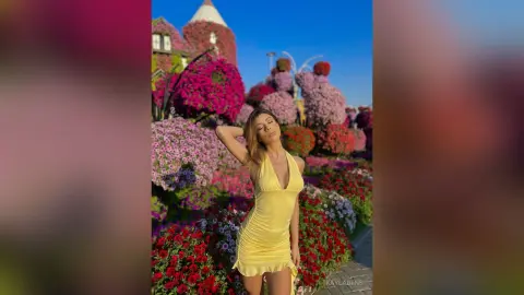 Stylish woman in a yellow sundress posing in a vibrant blooming flower garden under a bright sunny sky.