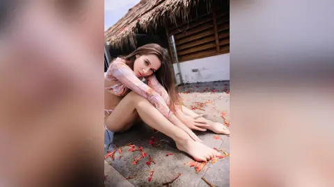 StacyHarland sits on a stone floor wearing a pink patterned long-sleeve crop top and matching bottoms, leaning forward with her hands resting near her feet.