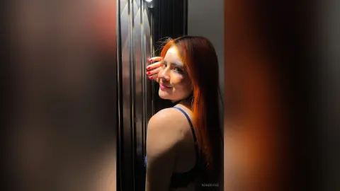 Redheaded woman in a blue lace bra posing in warm, dramatic sunlight by a dark window.