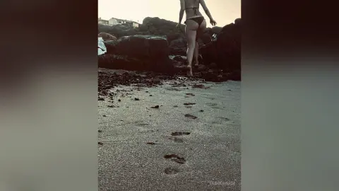 Slim woman in a black bikini walking on a sandy beach toward dark coastal rocks under a bright sky.