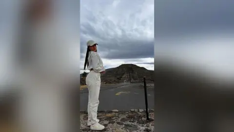 Woman in white casual outfit and cap standing against a cloudy sky at an ancient stone ruin site.