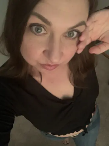 DirtyDaisyLou looks into the camera with a playful expression, wearing a black top and blue jeans.