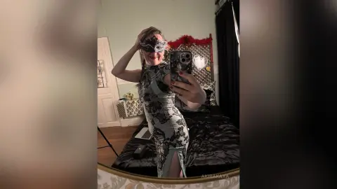 Smiling woman in a lace patterned dress and masquerade mask taking a mirror selfie in a cozy bedroom.