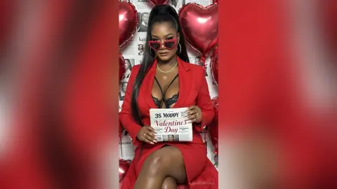 Stylish woman in red blazer and sunglasses holding a Valentine's Day newspaper against a romantic red backdrop.
