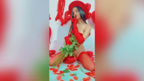 AmeliaStell poses naked in a red hat and red gloves, holding a long-stemmed rose against her chest while kneeling on red rose petals.