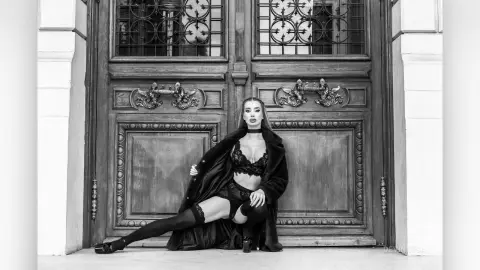 DeviNessa poses in black lace lingerie and a long dark coat while sitting in front of a large, ornate wooden door.