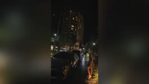 BiankaPerese stands outside at night next to a dark car, wearing a brown outfit that shows off her legs and curves.