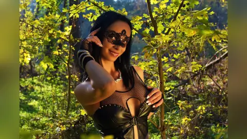 Mysterious woman in a black lace mask and leather outfit posing in a sunlit green forest.