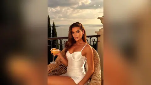 Glamorous woman in a white satin mini dress holding a cocktail on a luxury seaside balcony at sunset.