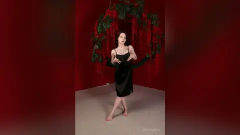 Elegant woman in a black slip dress posing against a moody red backdrop with hanging floral vines.