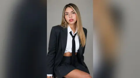 Young woman in a professional black blazer and white shirt with a black tie posing against a neutral background.