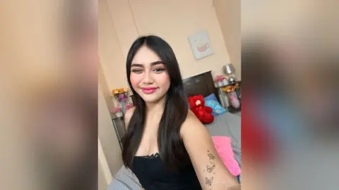 Smiling young woman in a black top winking in a cozy bedroom setting with a playful mood.