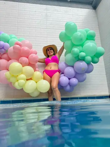 Smiling woman in pink bikini and sun hat posing by colorful pastel balloon arches next to a swimming pool.