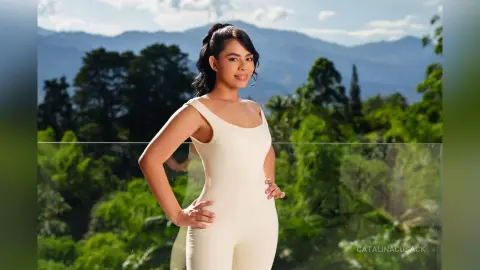 CatalinaCusack poses outdoors in a white tank-style bodysuit against a lush green forest and mountain backdrop.