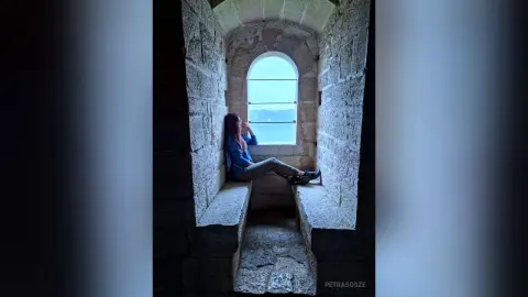 Melancholic woman in blue sweater sitting in a stone castle window overlooking a misty landscape.