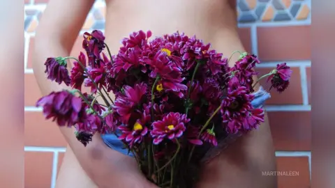 Woman in denim shorts holding a large bouquet of purple chrysanthemums against a brick wall background.