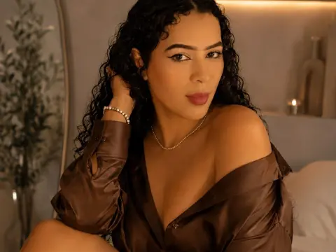 paulinaa_rosee poses with her long curly hair and a brown satin shirt falling off her shoulder, wearing a pearl necklace.