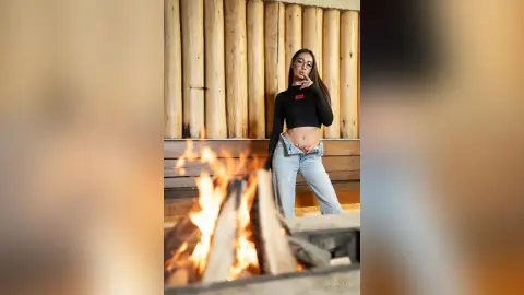 Young woman in black long-sleeve top and light jeans posing by a cozy outdoor campfire at a log cabin.