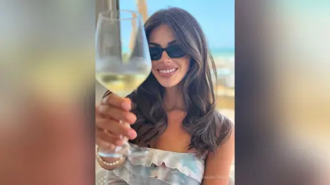AnaMathers smiles while holding a glass of white wine, wearing sunglasses and a light floral top with her long brown hair down.