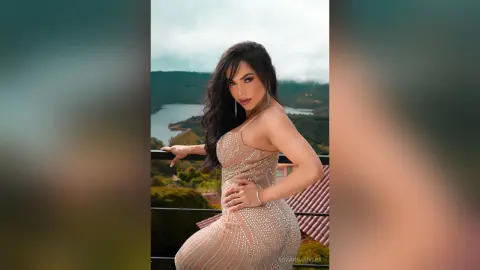 RoxannaRivera poses outdoors in a tight, sparkling tan dress, showing off her curves against a scenic lake and mountain background.