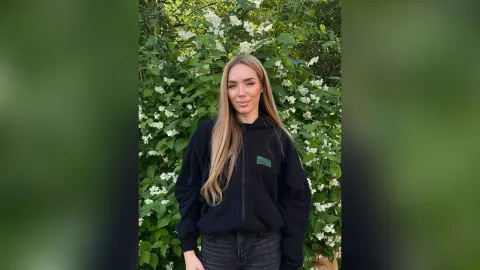 AmiliRay — AmiliRay stands in front of green bushes and white flowers wearing a black zip-up hoodie and dark jeans.