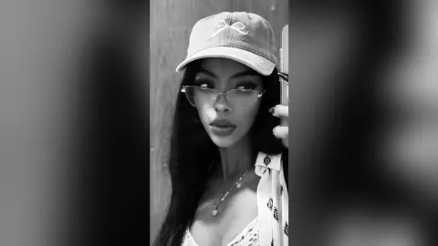 Young woman in a baseball cap and glasses posing for a moody black and white mirror selfie.