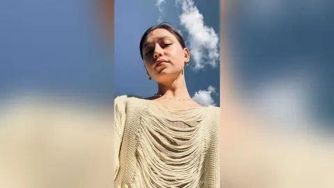RitaMoonllight looks up at the camera wearing a cream-colored fringed top against a blue sky with white clouds.